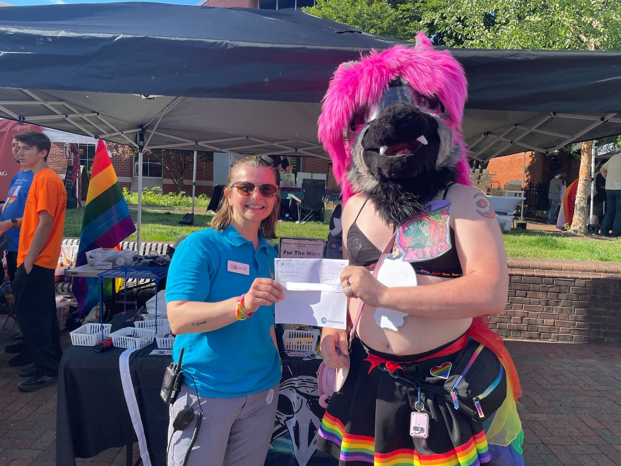 A person representing The Frederick Center holds up a check. They are wearing sunglasses and shorts and a blue shirt. There is a radio hanging from their belt. The other end of the check is held by our con chair Kitra. She is wearing a fursuit head with dark fur and bright purple hair. She is wearing a sport top and a skirt with rainbow colors on the hem.
