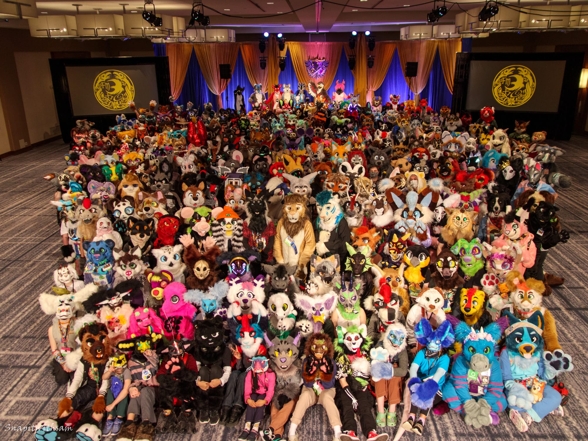 A large gathering of fursuiters sit and stand in a group on the floor at Fur the More 2024. They are colorful and many types and species. In the background the stage is draped in blue and gold. The two panels beside the stage have our gold fur the more circular raven logo